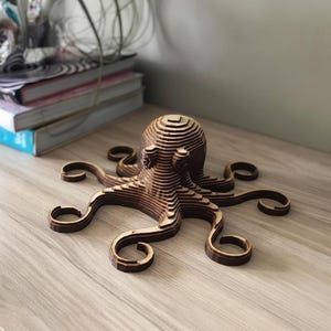 May include: A wooden octopus sculpture with a layered design. The sculpture is made of light and dark brown wood, with each layer visible. The octopus has eight curled tentacles and a rounded head. The sculpture is displayed on a light-coloured wooden surface.