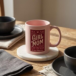 Puede incluir: Una taza de cer&aacute;mica rosa con las palabras "GIRL MOM" en un dise&ntilde;o decorativo. La taza tiene un asa y se apoya en un posavasos de m&aacute;rmol blanco. La taza tambi&eacute;n tiene el texto "EST. 2026 - GENUINE ARTICLE".