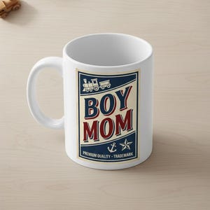 Puede incluir: Taza de cer&aacute;mica blanca con un gr&aacute;fico azul, rojo y crema que dice "BOY MOM". El dise&ntilde;o incluye un tren, un ancla y una estrella. Tambi&eacute;n se incluye el texto "PREMIUM QUALITY - TRADEMARK".