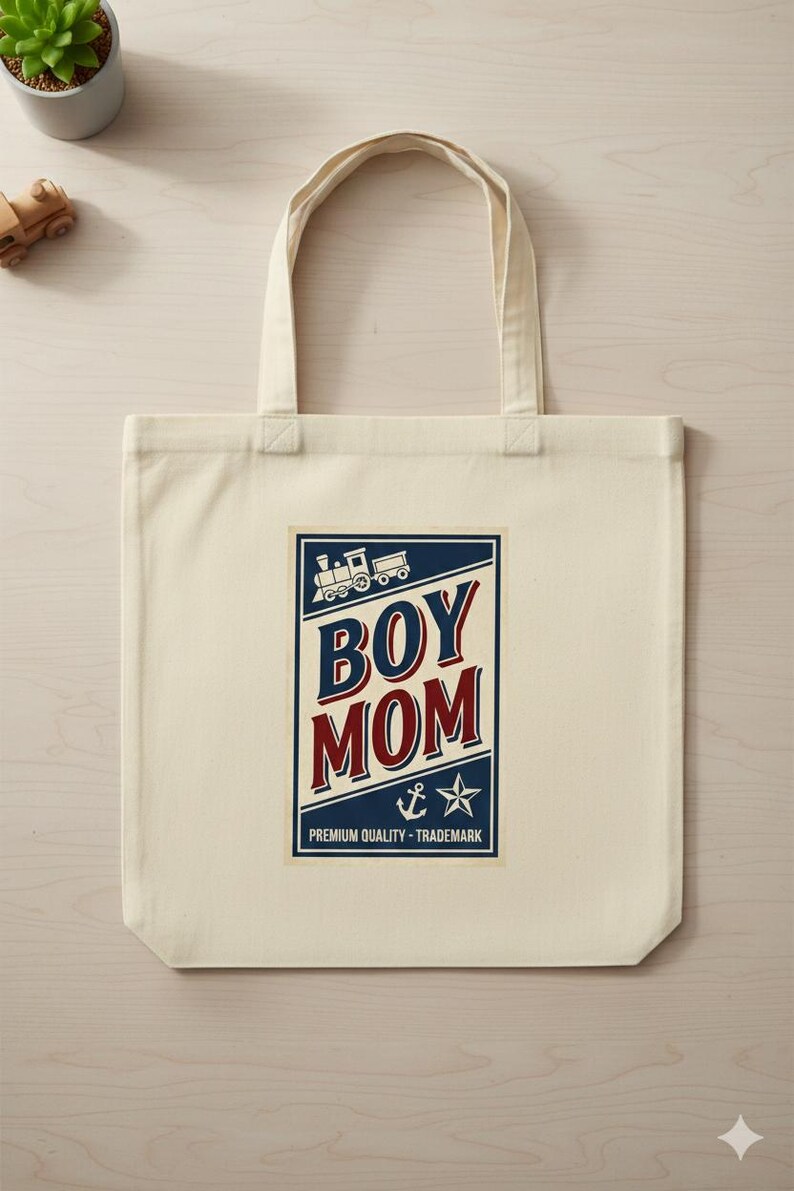 Puede incluir: Bolso tote de color crema con un gr&aacute;fico rectangular. El gr&aacute;fico presenta las palabras "BOY MOM" en rojo y azul, una ilustraci&oacute;n de tren, un ancla y una estrella. Tambi&eacute;n se incluye el texto "PREMIUM QUALITY - TRADEMARK".