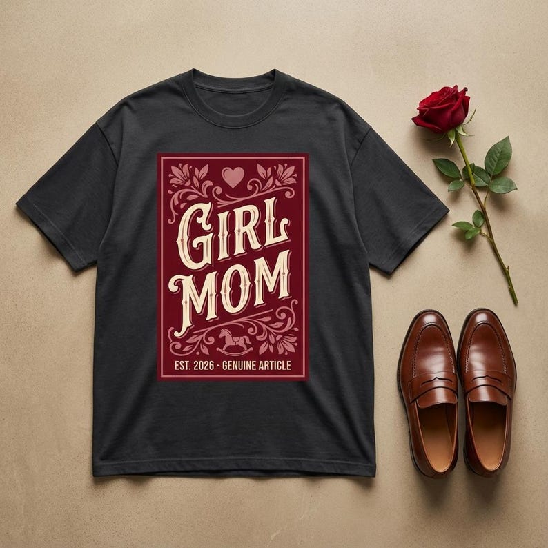 Puede incluir: Camiseta negra con un gr&aacute;fico granate que dice "GIRL MOM" en un estilo vintage. El gr&aacute;fico incluye un coraz&oacute;n, elementos florales y el texto "EST. 2026 - GENUINE ARTICLE". Una rosa roja y mocasines de cuero marr&oacute;n tambi&eacute;n est&aacute;n en la imagen.