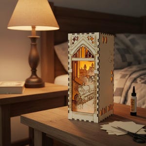 May include: A wooden, rectangular night light with a detailed laser-cut design. The light box features a train, castle, and arched window design. The light is on, illuminating the interior scene. The night light is on a wooden table next to a lamp.