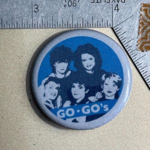 May include: A blue and white button with a picture of the band Go-Go's. The band members are all women and are wearing black clothing. The text "GO-GO'S" is printed in white on a blue background.