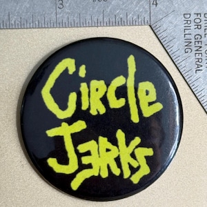 May include: A black and yellow round button with the text "Circle Jerks" in a yellow, handwritten font.
