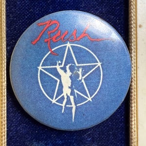 May include: A round, blue button featuring the band name "Rush" in red script. A white silhouette of a figure within a star is centered on the button, a classic music collectible.
