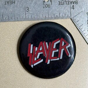 May include: A black button with a red and white Slayer logo. The logo is a stylized font with the word "Slayer" in red and white.