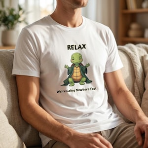 Może przedstawiać: Biała koszulka z kreskówkowym żółwiem w zrelaksowanej pozie, w zielonej szacie. Słowo "RELAX" znajduje się nad żółwiem, a fraza "We're Going Nowhere Fast" poniżej. Koszulka jest wykonana z miękkiego materiału.