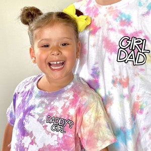 May include: A father and daughter wearing matching tie-dye shirts. The father's shirt says "Girl Dad" and the daughter's shirt says "Daddy's Girl".