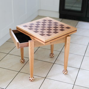 May include: A wooden chess table with a checkerboard top and a drawer. The table has four legs with ball-shaped feet.