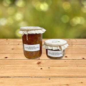 May include: Two glass jars of chow chow relish, one large and one small, are displayed on a wooden surface. The jars are sealed with fabric and twine. Each jar has a label with the text "Chow Chow Relish" and a list of ingredients.