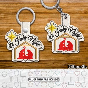 May include: Two keychains with a religious theme. Each keychain features a cream-colored leatherette tag with a stitched design of a nativity scene in red and gold, with the words "Holy Night!" in black script. The keychains have silver-colored key rings.