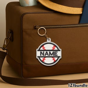 May include: A baseball-themed keychain in white and red, featuring the word "NAME" in black. The keychain is attached to a brown bag with a zip. The bag has a shoulder strap and a gold-coloured zip.
