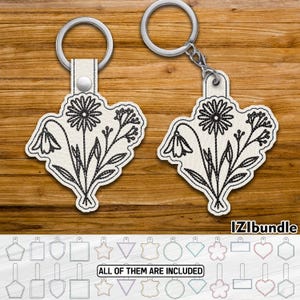 Könnte beinhalten: Zwei cremefarbene Schlüsselanhänger mit floralem Design, die eine Gänseblümchen und andere Blumen zeigen. Die Blumen sind mit schwarzem Faden gestickt. Jeder Schlüsselanhänger hat einen silbernen Ring und einen Lederriemen. Der Text "IZIbundle" und "ALL OF THEM ARE INCLUDED" sind ebenfalls sichtbar.