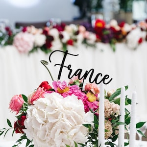 Può includere: Una tavola bianca apparecchiata con un centrotavola di ortensie bianche, fiori rosa e rossi e verde. Ci sono tre candele bianche in candelabri dorati. La tavola è apparecchiata con piatti bianchi, tovaglioli rossi e bicchieri da vino. Un cartello nero con la parola "Francia" è posizionato al centro dell'arrangiamento.
