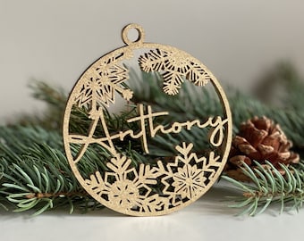 May include: A gold wooden Christmas bauble with a snowflake design and the name "Anthony" written in cursive.