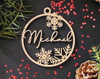 May include: A wooden Christmas bauble featuring a snowflake design and the name "Michael" in a cursive script. The bauble is surrounded by red glitter against a black backdrop.