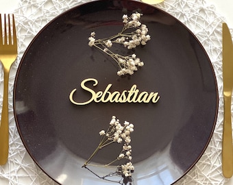 Puede incluir: Un plato negro con un mantel individual de encaje blanco, cubiertos dorados y una tarjeta de nombre dorada con el nombre "Sebastian" en ella. El plato está decorado con pequeñas flores blancas.