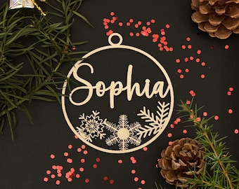 May include: A wooden Christmas bauble with the name "Sophia" written in cursive. The bauble is decorated with snowflakes and pine branches. It is surrounded by red glitter on a black background.