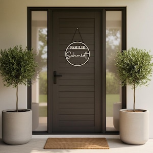 Könnte beinhalten: Eine dunkelgraue Haustür mit einem kreisförmigen weißen Schild mit den Worten "FAMILIE" und "Schmidt". Zwei Topf-Olivenbäume stehen zu beiden Seiten der Tür. Eine braune Fußmatte liegt auf dem Boden.