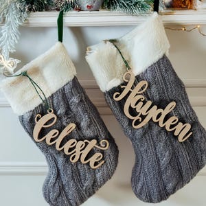 May include: Two gray cable-knit Christmas stockings with white faux fur cuffs hang from a white mantel. Wooden name tags that read "Celeste" and "Hayden" are attached to the stockings with green ribbon. Festive holiday decor.