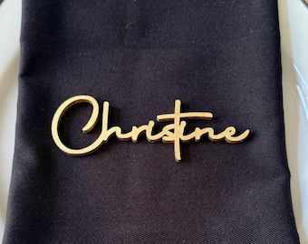 May include: A gold-colored nameplate with the name "Christine" in a cursive font. The nameplate is placed on a black napkin, suggesting a place setting for an event or dinner.