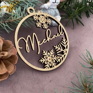 Pu&ograve; includere: Un ornamento natalizio in legno con il nome "Michael" scritto in corsivo. L'ornamento &egrave; decorato con fiocchi di neve e rami di pino.