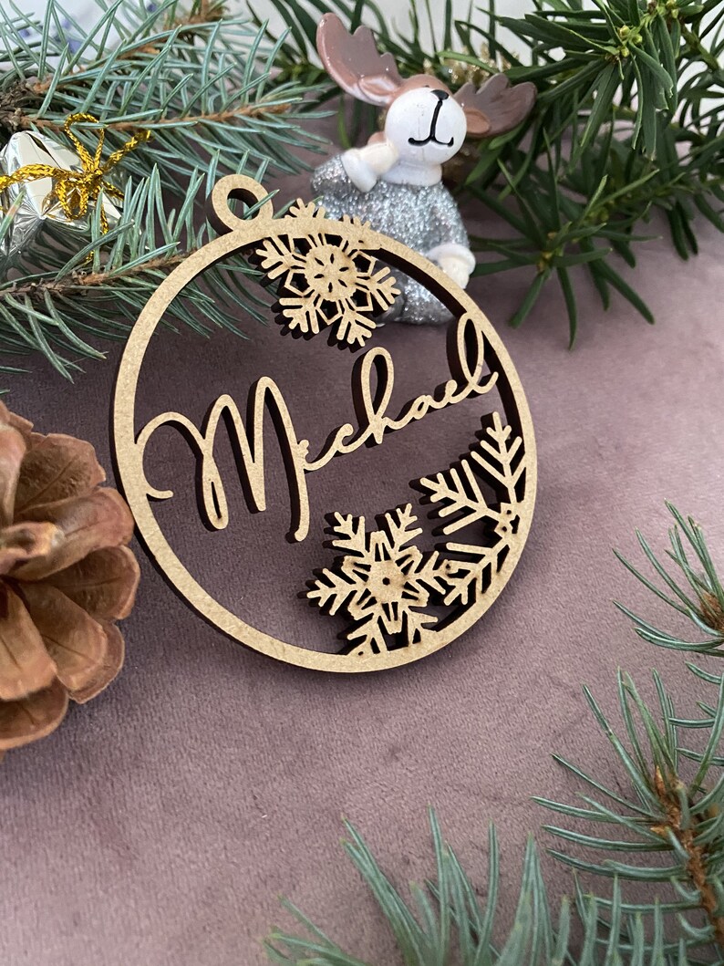 Pu&ograve; includere: Un ornamento natalizio in legno con il nome "Michael" scritto in corsivo. L'ornamento &egrave; decorato con fiocchi di neve e rami di pino.