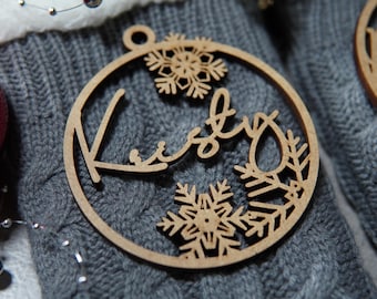 May include: A wooden Christmas bauble with the name "Kirby" cut out in a cursive font, surrounded by snowflake designs. The bauble is circular with a hole at the top for hanging. The background is a grey knitted fabric.