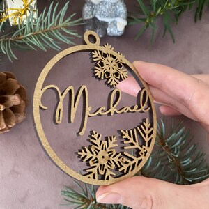 Pu&ograve; includere: Un ornamento natalizio rotondo in legno con il nome "Michael" scritto in corsivo. L'ornamento &egrave; decorato con fiocchi di neve ritagliati nel legno.
