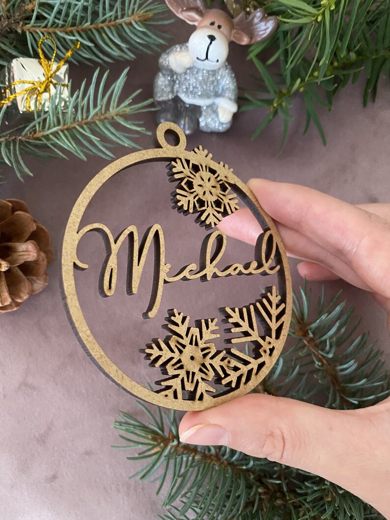 Pu&ograve; includere: Un ornamento natalizio rotondo in legno con il nome "Michael" scritto in corsivo. L'ornamento &egrave; decorato con fiocchi di neve ritagliati nel legno.