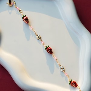 Peut inclure: Un bracelet délicat en chaîne dorée avec des perles roses et blanches, orné de petits charms en forme de fraises et de fleurs.