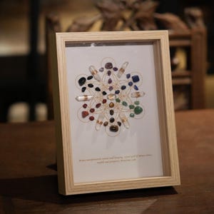 May include: A framed crystal grid with a variety of colored stones and clear quartz crystals. The grid is surrounded by a gold wire and the text "Money manifestation crystal wall hanging, crystal grid of money, wealth and prosperity abundance grid" is printed below the grid.