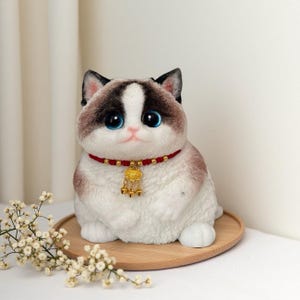 Könnte beinhalten: Plüschkatzenfigur mit weißem Körper, braunen und schwarzen Markierungen und großen blauen Augen. Sie trägt ein rotes Halsband mit einem goldenen Anhänger und sitzt auf einem runden Holztablett. Die Katze ist ein Dekorationsartikel.