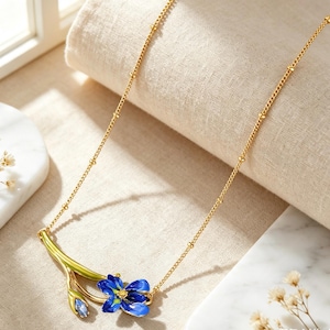 May include: A gold-tone chain necklace featuring a pendant shaped like a flower stem with a blue enamelled blossom and a small blue bud. The metal stem is finished in a bright green enamel, and the chain includes small gold beads at intervals.