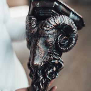 May include: A decorative shelf bracket made of metal in the shape of a ram's head. The bracket is painted in a dark brown color with a copper finish. The ram's head has detailed features, including horns, eyes, and a beard.
