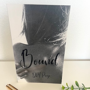 May include: A black and white photo of a person's back with the word "Bound" in black cursive lettering and the author's name "DM Page" in black lettering below.
