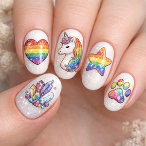 Pode incluir: Uma mão com cinco unhas pintadas, cada uma com um design diferente. Os desenhos incluem um coração arco-íris, um unicórnio com uma crina arco-íris, uma estrela arco-íris, cristais coloridos e uma pegada arco-íris. As unhas têm uma base branca brilhante.