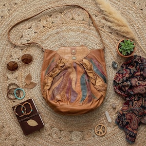 May include: A brown leather shoulder bag with colorful leaf-like accents, a textured flap, and a long strap. The image also includes sunglasses, bracelets, a journal, a scarf, and a small potted plant, all arranged on a woven rug.