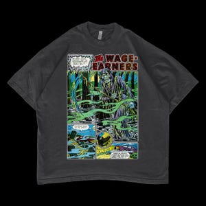 Op de afbeelding: Een donkergrijs T-shirt met een stripboekgrafiek van een grim reaper in een moeras. Het stripboekpaneel luidt "The Wage-Earners".