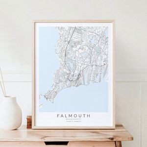 May include: A framed map print of Falmouth, Massachusetts, with a light blue background and black lines showing the streets and roads. The map is on a light wood table with a white vase and a dried grass plant.