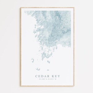 May include: A framed print of a blue watercolor map of Cedar Key, Florida. The coordinates 29.1386° N, 83.0351° W are printed below the map.