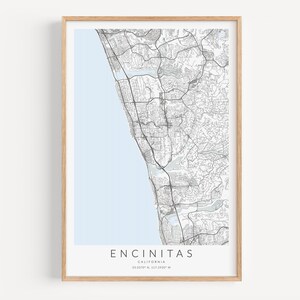 May include: A black and white map print of Encinitas, California, with a light blue background. The map shows the streets and roads of the city. The print is framed in a light wood frame.