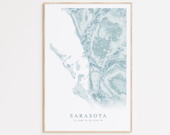 May include: A blue and white print of a map of Sarasota, Florida. The map is framed in a light wood frame. The text "Sarasota" and the coordinates "27.3467° N, 82.5310° W" are printed below the map.