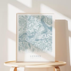 May include: A framed print of a map of London in a light blue color scheme. The map shows the Thames River winding through the city. The text "LONDON" is printed below the map, along with the coordinates "51.5072&deg; N, 0.1275&deg; W".