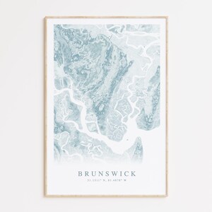 May include: A framed print of a blue and white watercolor map of Brunswick, Georgia. The map shows the waterways and islands of the area. The text "BRUNSWICK 31.1317° N, 81.4879° W" is printed below the map.