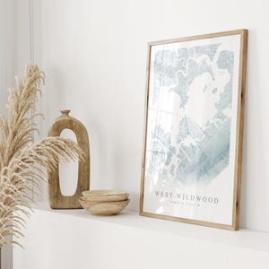May include: A framed print with a light blue and white abstract design, labeled "WEST WILDWOOD". The print is in a light wood frame, displayed with wooden bowls, a decorative wooden sculpture, and dried pampas grass. The overall aesthetic is minimalist and natural.