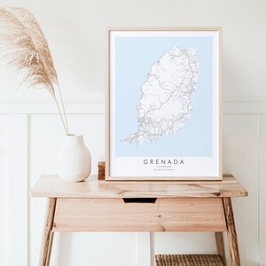 May include: A framed map of Grenada, a Caribbean island, with a light blue background and white lines representing roads. The frame is light wood and the map is on a light wood table with a white vase and a dried plant.