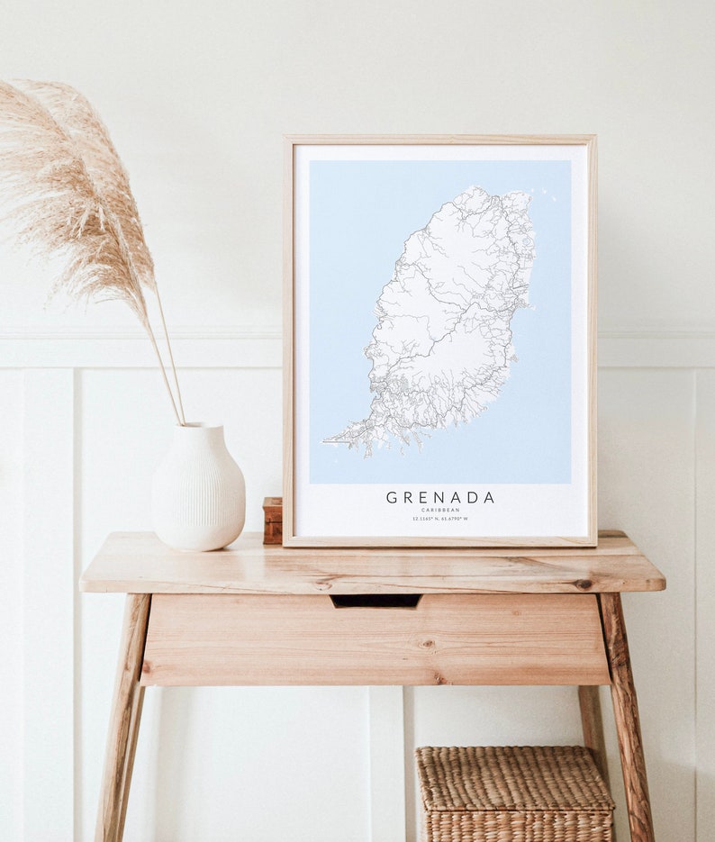 May include: A framed map of Grenada, a Caribbean island, with a light blue background and white lines representing roads. The frame is light wood and the map is on a light wood table with a white vase and a dried plant.