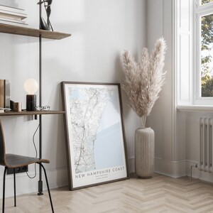 K&ouml;nnte beinhalten: Ein gerahmter Kartenabdruck der K&uuml;ste von New Hampshire mit hellblauem Hintergrund und schwarzen Linien. Der Druck h&auml;ngt an einer wei&szlig;en Wand in einem Heimb&uuml;ro. Eine Vase mit getrocknetem Pampasgras steht auf dem Boden neben dem Druck.