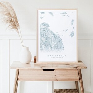 May include: A framed print of a blue and white map of Bar Harbor, Maine. The map is in a light wood frame and is sitting on a wooden table.